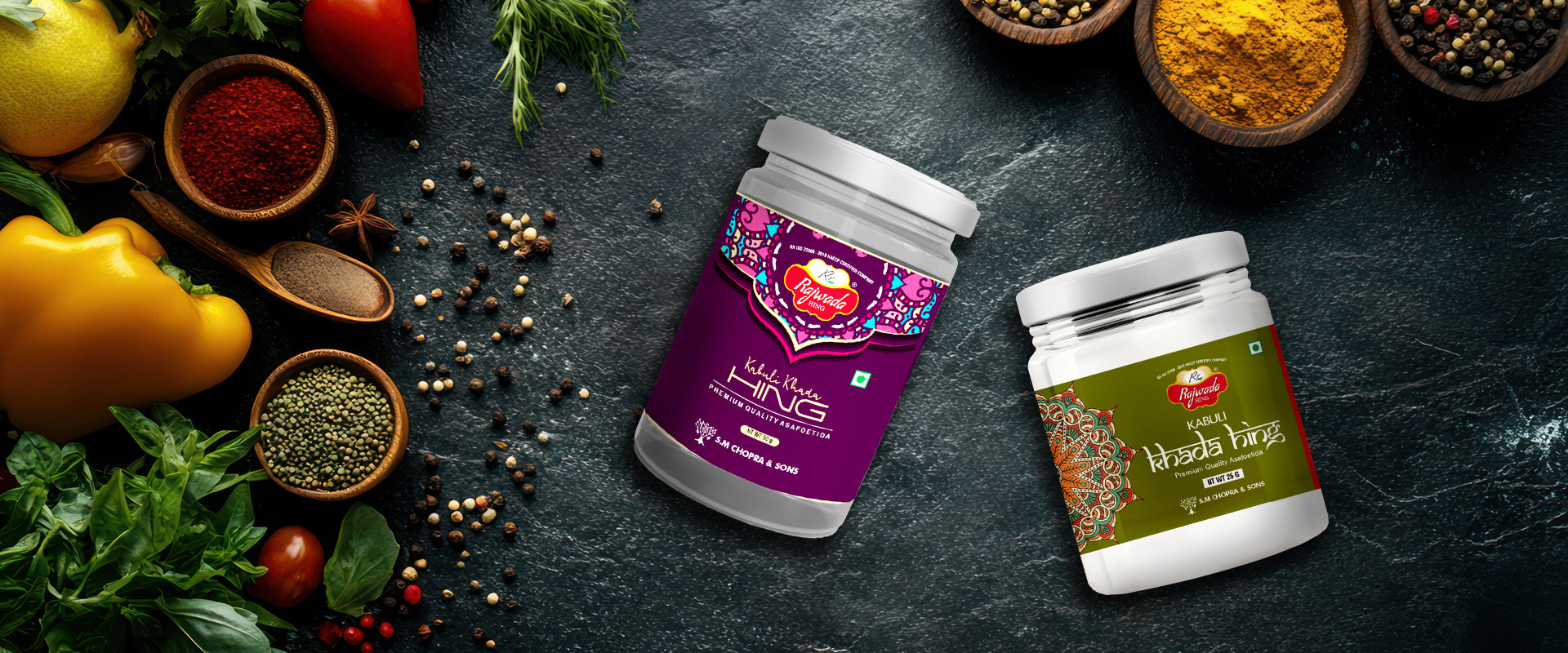 Two Rajwada Hing jars with colorful labels on a dark surface with various spices and herbs.