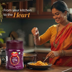 Woman in traditional attire adding Sanyukt Bandhani Hing Powder – Premium Quality for Perfect Tadka to food with a purple spice jar labeled 'Panchagavya' in a kitchen setting.